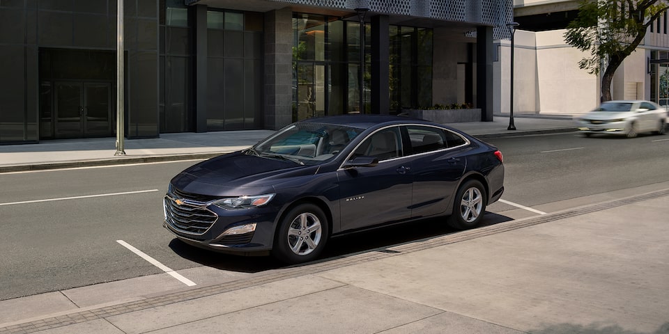 Side profile view of the 2024 Chevrolet Malibu parked in front of a modern building.