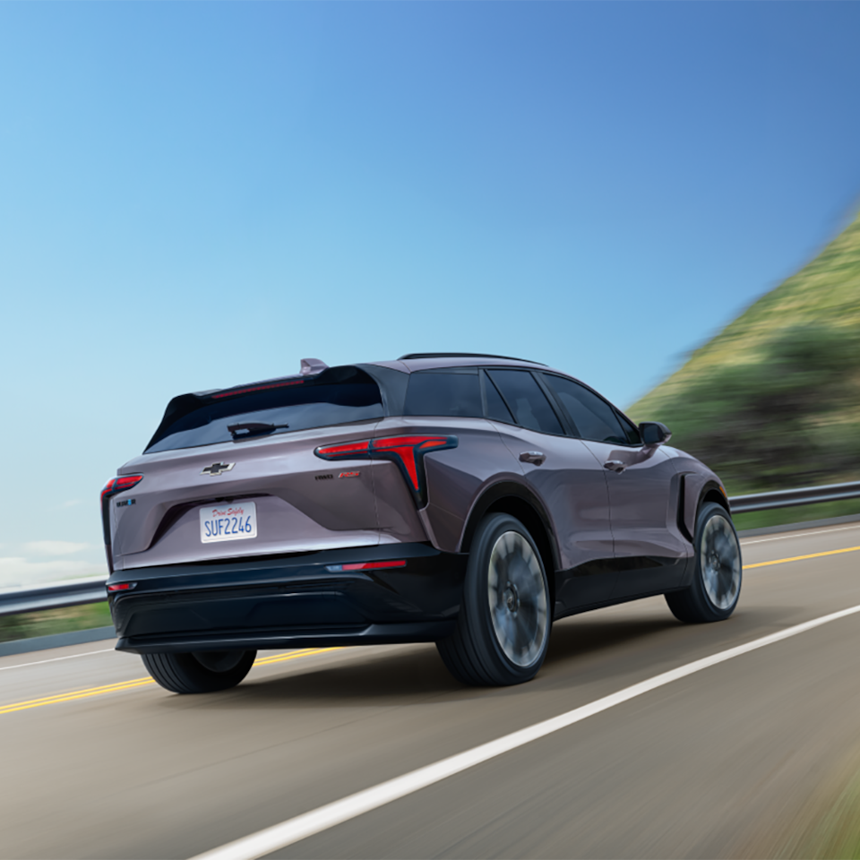 Rear three quarters view of the 2025 Chevrolet Blazer EV driving down the road under a clear blue sky.