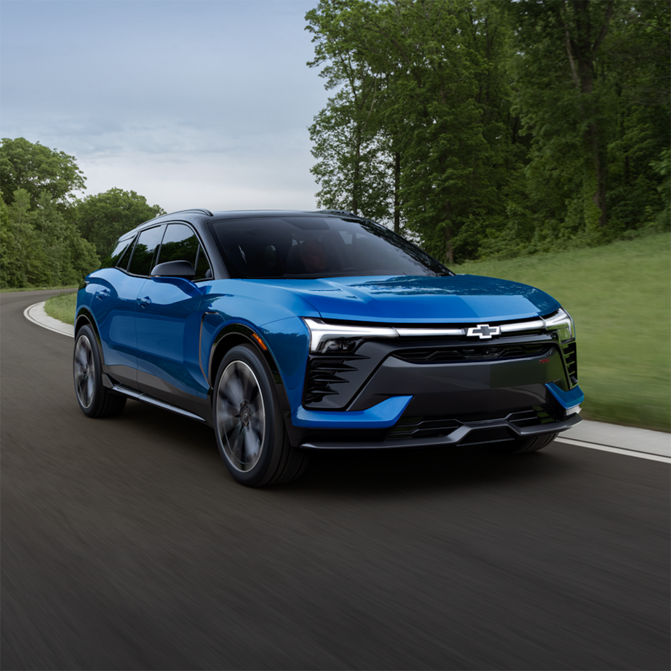 Three quarters view of a 2025 Chevrolet Blazer EV driving next to grassy plain and trees.