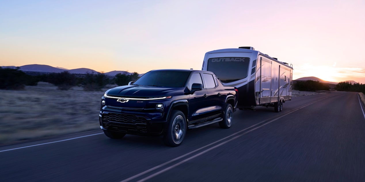 Front-side view of a 2025 Chevrolet Silverado EV with a trailer behind on a road at evening.