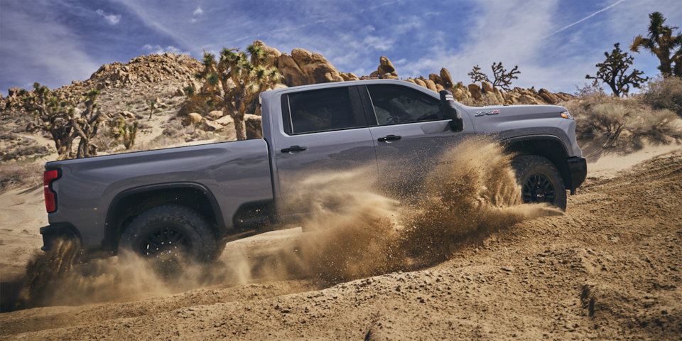 Side view of the 2025 Chevrolet Silverado HD pickup truck driving past a rough terrain.