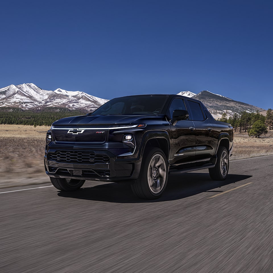 Three-quarters view of the front end of the 2024 Chevrolet Silverado EV driving along a forest-lined road.