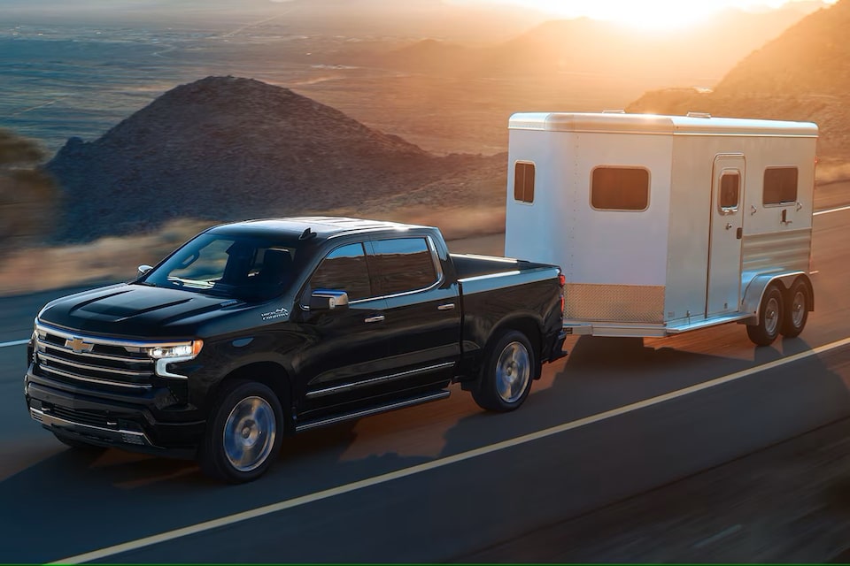 A black 2025 Chevrolet Silverado LD Work Truck pulling a trailer down the road on a sunny day.